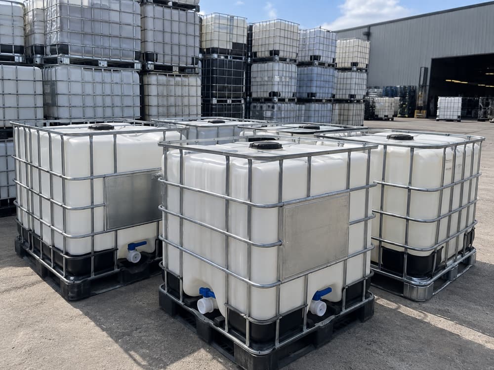 Rows of washed white IBC totes in steel cages on black plastic pallets, stacked two high in a sunlit yard