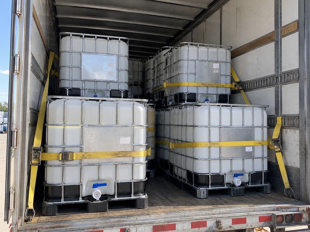 Four IBC totes strapped with yellow ratchet straps inside a trailer, valves facing inboard